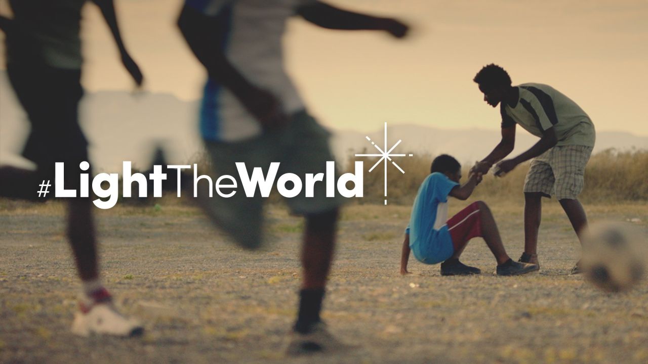 En dreng hjælper en anden dreng med at stå op efter at være faldet ved at spille fodbold fra videoen Oplys verden